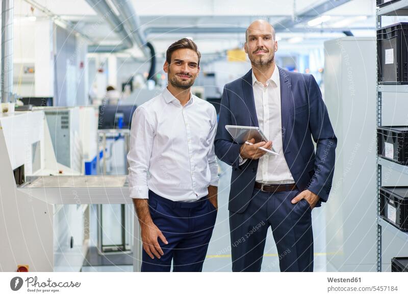 Confident mature businessman holding digital tablet while standing by young male engineer at factory color image colour image indoors indoor shot indoor shots