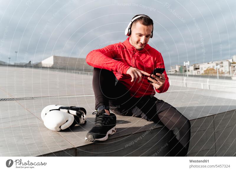 Sportsman wearing headphones using mobile phone while sitting by boxing gloves outdoors color image colour image location shots outdoor shot outdoor shots day