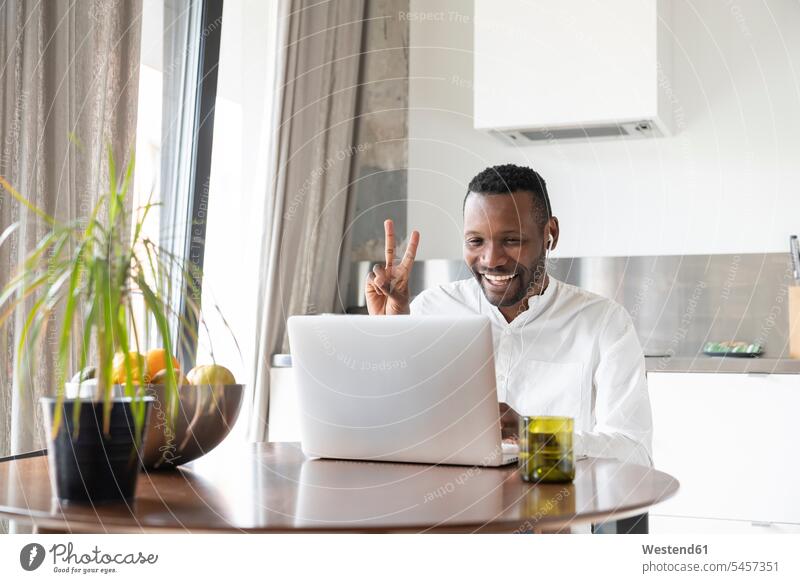 Portrait of happy man sitting at kitchen table during video chat showing victory sign computers Laptop Computer Laptop Computers laptops notebook Seated delight