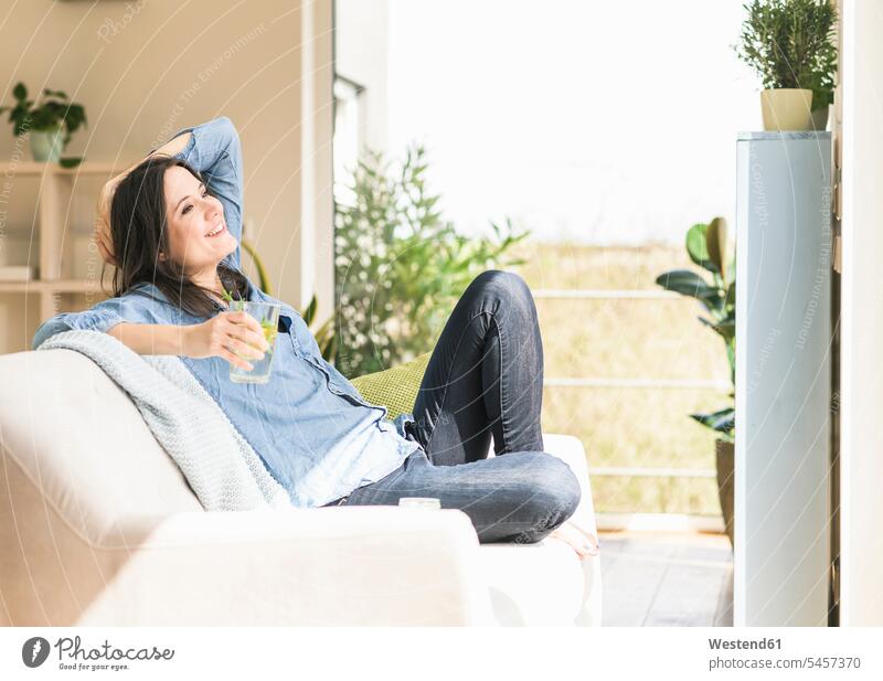 Happy woman with a glass of infused water sitting on the couch at home females women Seated Water settee sofa sofas couches settees Glass Drinking Glasses
