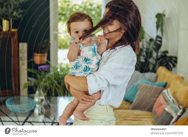 Baby girl pulling mother’s hair in living room daughter daughters mommy mothers ma mummy mama baby infants nurselings babies child children family families