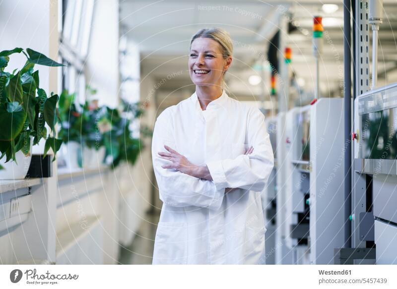 Confident mature female scientist with hands in lab coat pockets standing by machinery at laboratory color image colour image indoors indoor shot indoor shots