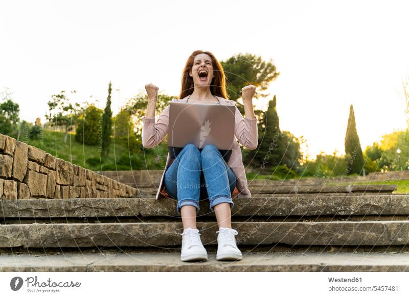 Young redheaded woman using laptop and cheering, sitting on steps in a park human human being human beings humans person persons vanquisher winner types winners