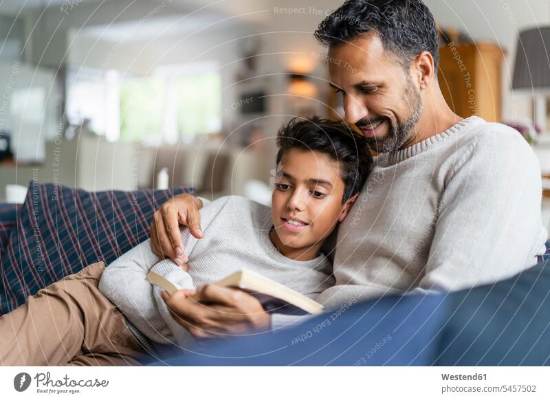 Father lying with son on couch in living room reading book books jumper sweater Sweaters couches settee settees sofa sofas cuddle snuggle snuggling smile Seated