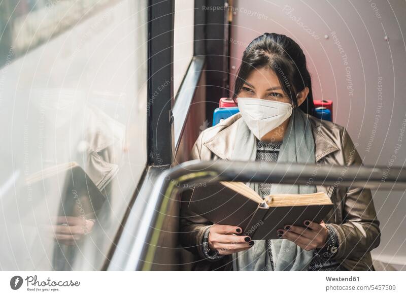Woman wearing protective mask, looking through window while reading book in bus color image colour image Vehicle Interior Selective focus Differential Focus
