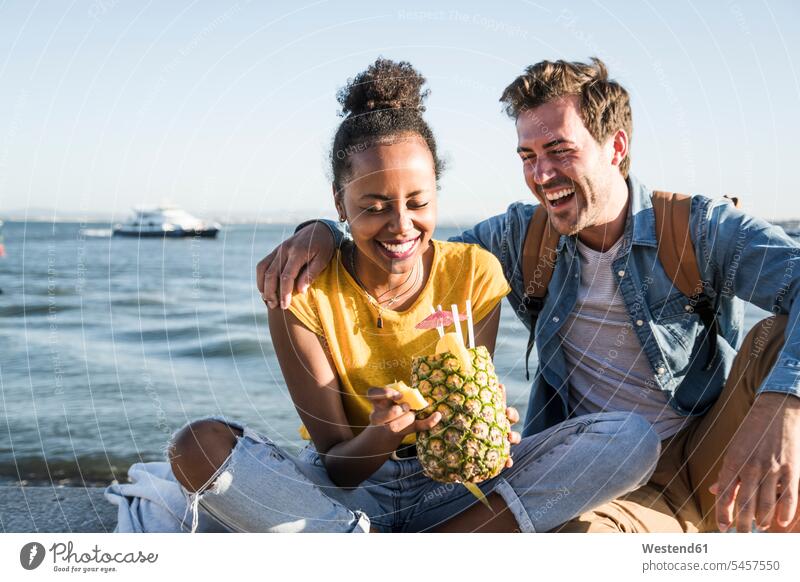 Happy young couple sitting on pier at the waterfront with a pineapple, Lisbon, Portugal human human being human beings humans person persons African black