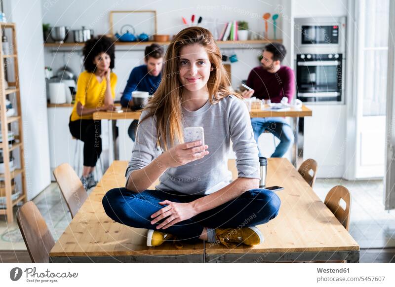 Woman with cell phone sitting on dining table at home with friends in background human human being human beings humans person persons caucasian appearance