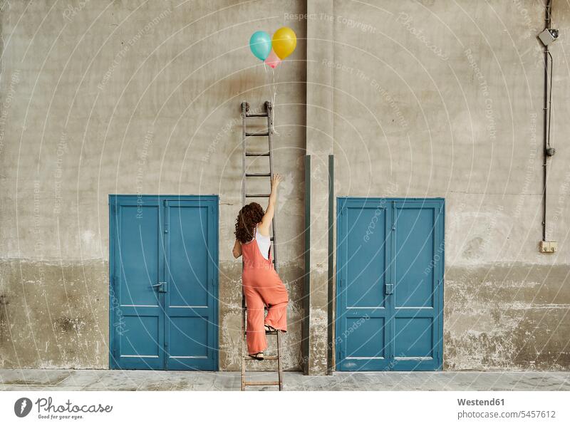 Woman climbing ladder leaning on wall while reaching for colorful helium balloons color image colour image indoors indoor shot indoor shots interior