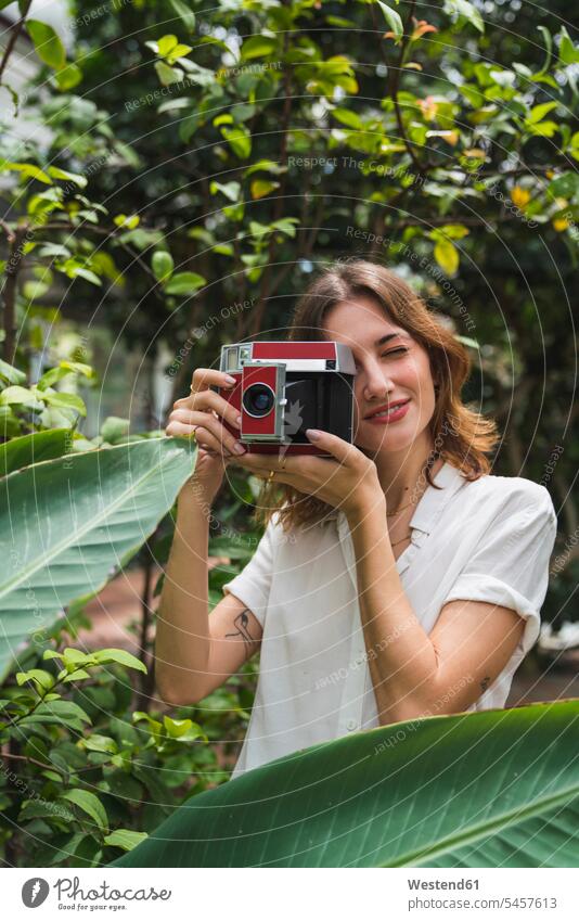 Young woman in greenhouse, taking pictures with an instant camera young women young woman photographing polaroid camera females Adults grown-ups grownups adult