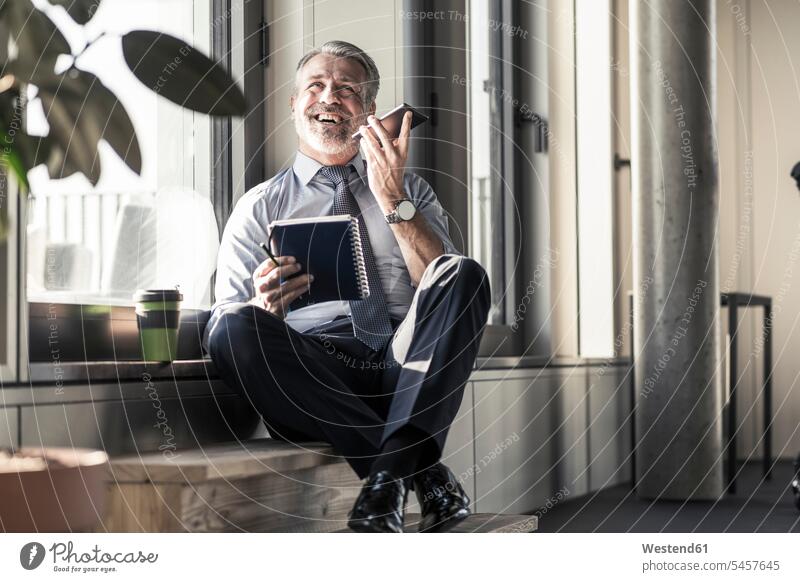 Happy mature businessman with notebook sitting at the window using cell phone on the phone call telephoning On The Telephone calling Seated notebooks happiness
