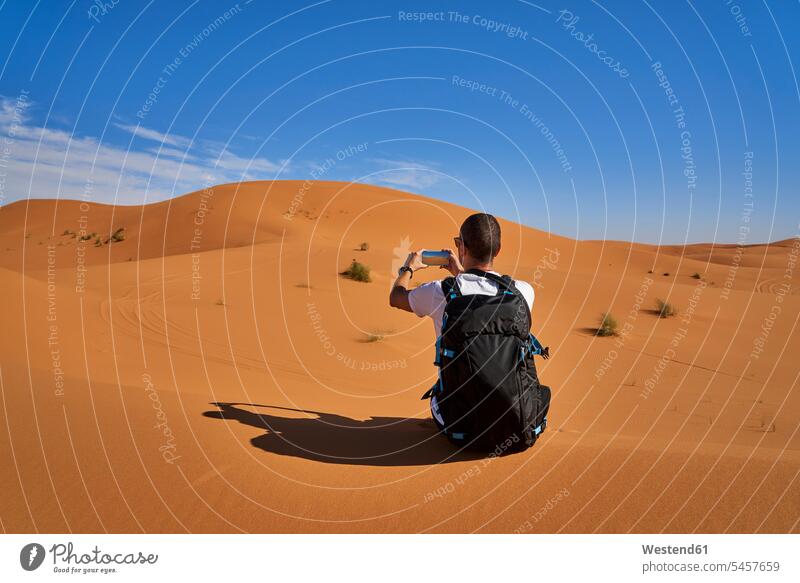 Morocco, man with backpack sitting on desert dune taking picture with smartphone Smartphone iPhone Smartphones photographing Seated desert dunes men males