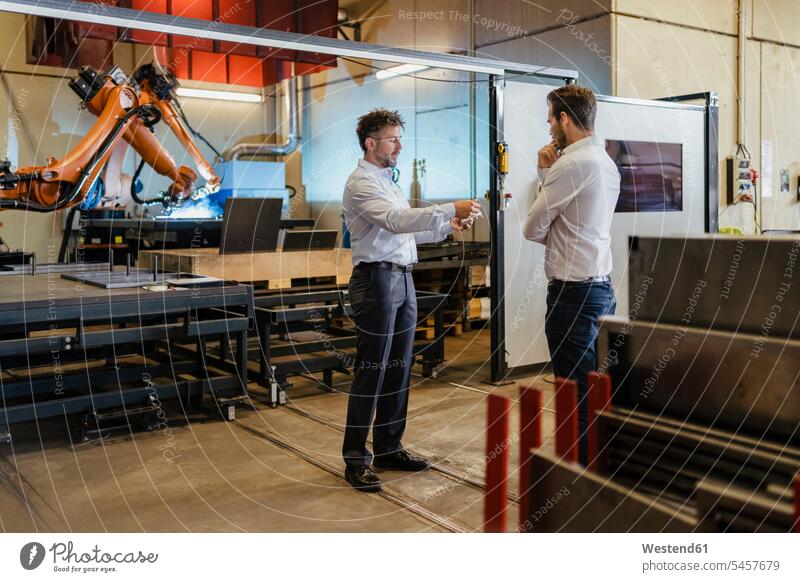 Young and mature businessman having discussion while standing against robotic machine at factory color image colour image indoors indoor shot indoor shots