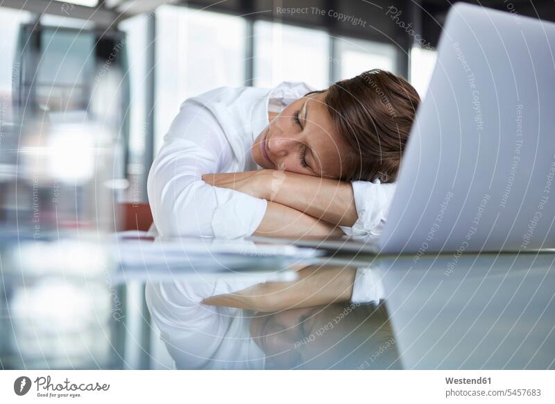 Businesswoman with closed eyes lying on glass table in office in front of laptop offices office room office rooms businesswoman businesswomen business woman