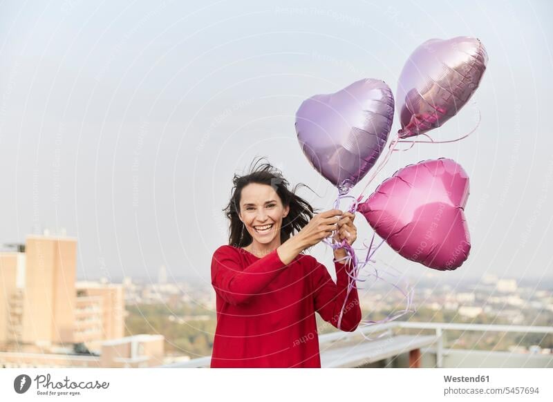 Smiling mature woman holding heart shape balloon while standing on building terrace color image colour image outdoors location shots outdoor shot outdoor shots