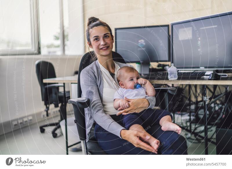 Working mother with baby on her lap, sitting in office working mother baby boys male babies infants people persons human being humans human beings Office Desk