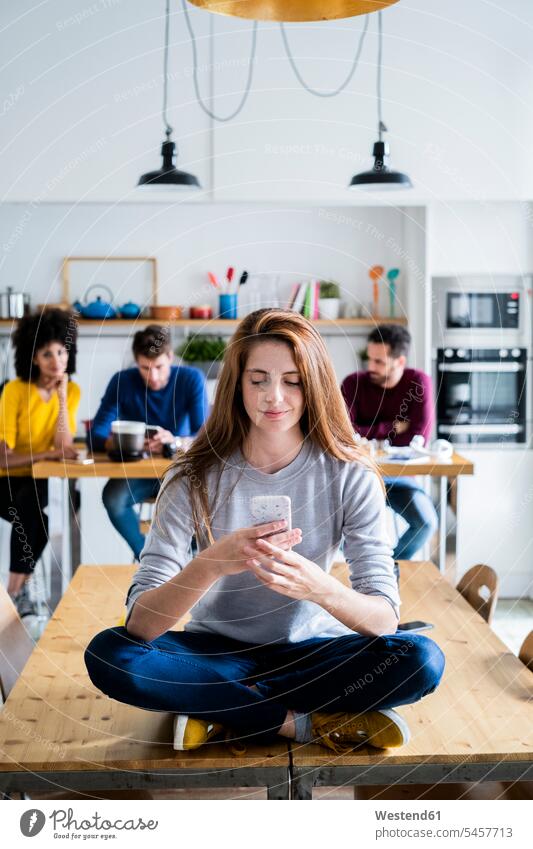 Woman with cell phone sitting on dining table at home with friends in background human human being human beings humans person persons caucasian appearance