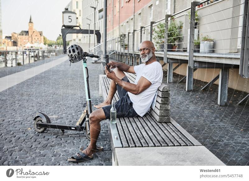 Portrait of mature man with E-Scooter relaxing on bench in summer using cell phone, Cologne, Germany Bottles Glass Bottles T- Shirt t-shirts tee-shirt benches