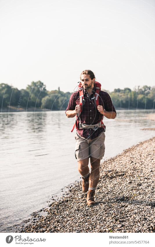 Young man with backpack walking at the riverside rucksacks backpacks back-packs River Rivers going men males riverbank water waters body of water Adults