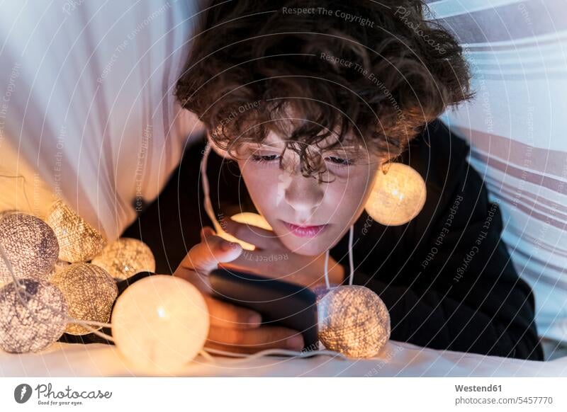 Boy with chain of lights underneath bedcover using cell phone human human being human beings humans person persons curl curled curls curly hair Bed - Furniture