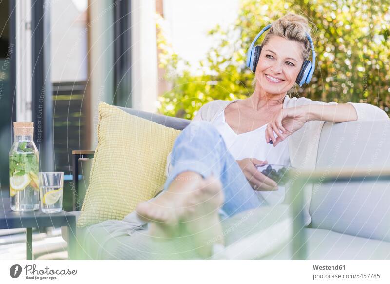 Portrait of relaxed mature woman sitting on terrace listening music with headphones while eating blueberries Bottles Glass Bottles Crockery Tableware
