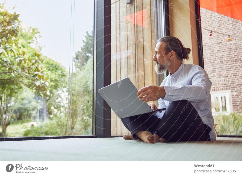 Bearded mature man with laptop looking through window while sitting in tiny house color image colour image Germany casual clothing casual wear leisure wear
