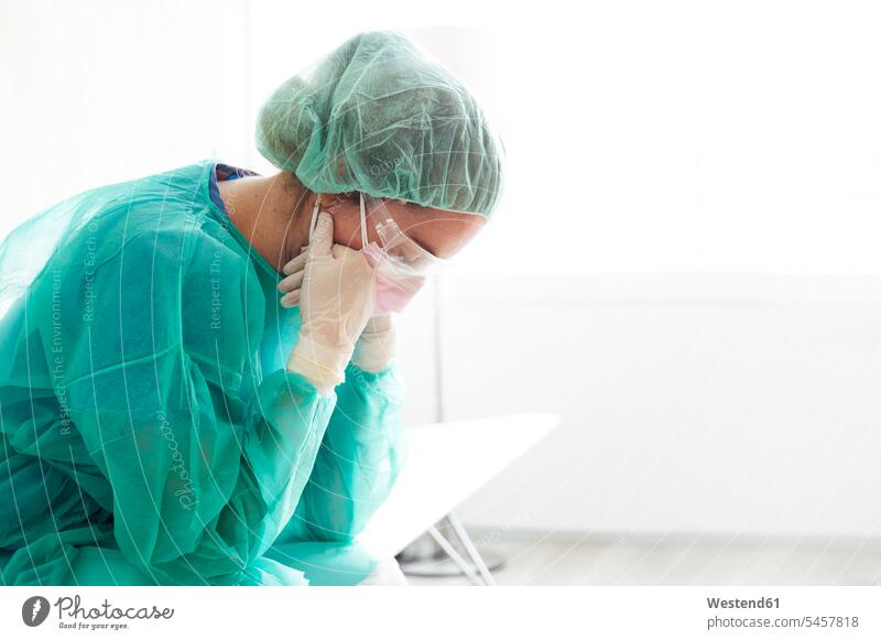 Sad doctor wearing protective workwear sitting against window in hospital color image colour image indoors indoor shot indoor shots interior interior view