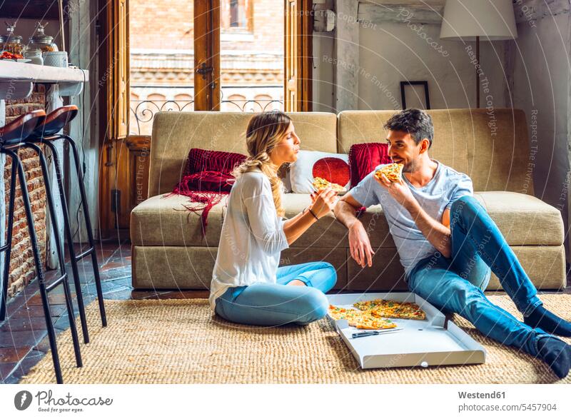 Young couple sitting on floor of living room and eating pizza from box boxes couches settee settees sofa sofas carpets rug rugs smile Seated delight enjoyment
