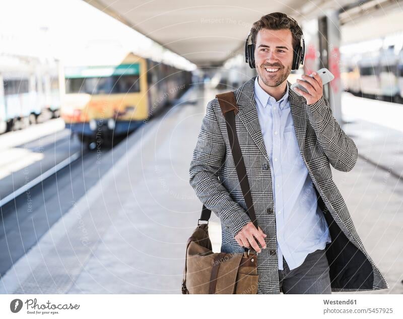 Smiling young businessman with cell phone and headphones at the train station human human being human beings humans person persons caucasian appearance