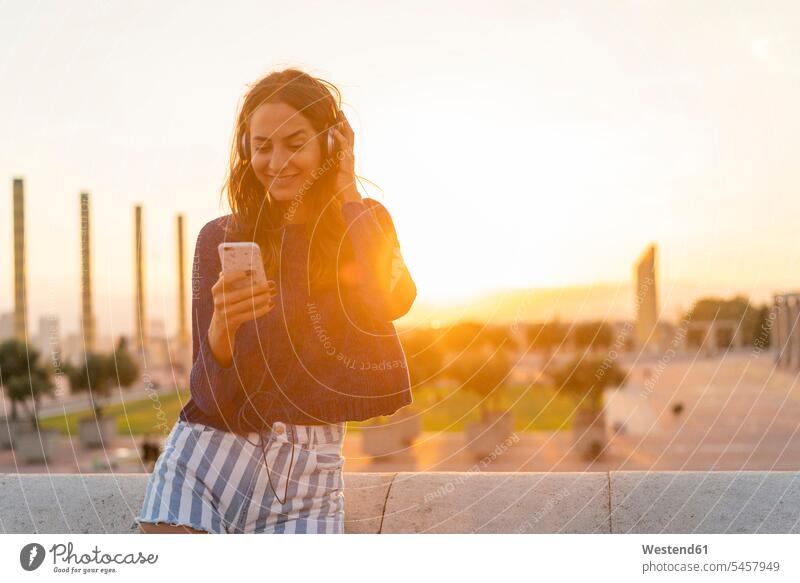 Spain, Barcelona, Montjuic, smiling young woman with cell phone and headphones at sunset mobile phone mobiles mobile phones Cellphone cell phones sunsets