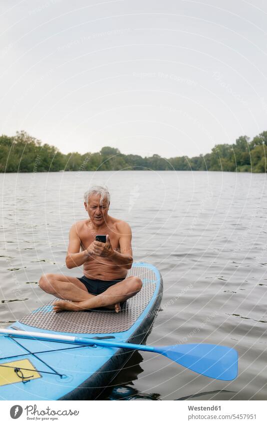 Senior man sitting on SUP board on a lake using cell phone men males lakes Paddleboard standup paddleboard paddle board Paddleboards senior men senior man