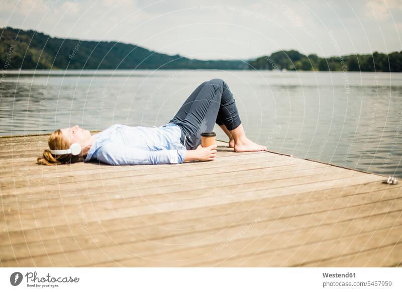 Woman lying on jetty at a lake with headphones and takeaway coffee headset jetties Coffee laying down lie lying down woman females women lakes Drink beverages
