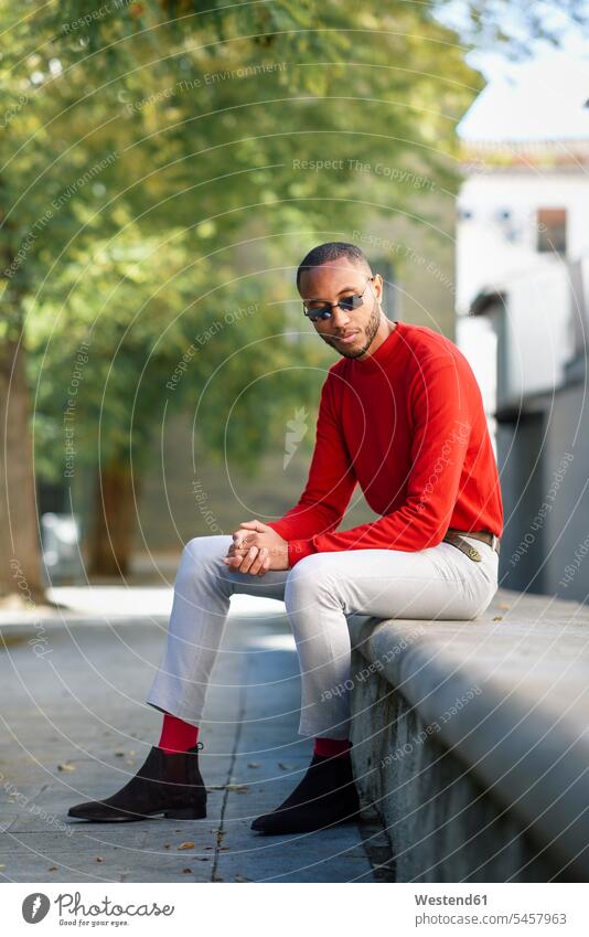 Portrait of fshionable young man wearing red pullover and sunglasses sitting on stone bench sweater jumper Sweaters fashionable men males portrait portraits