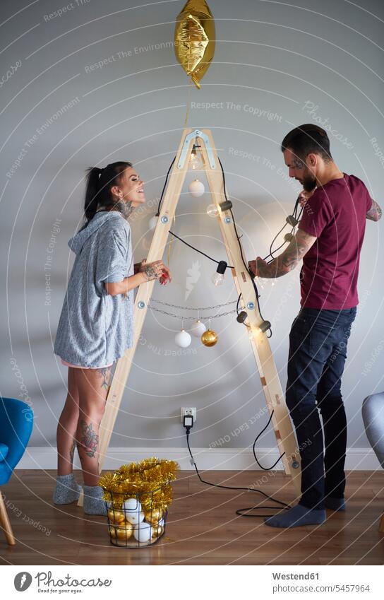 Modern couple decorating the home at Christmas time using ladder as Christmas tree X-Mas yule Xmas X mas at home twosomes partnership couples modern