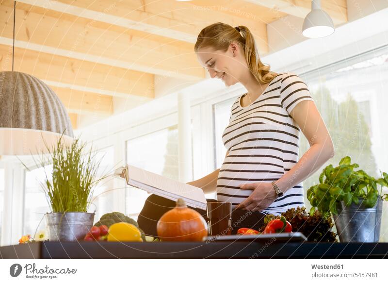 Smiling pregnant woman reading book in kitchen at home Pregnant Woman females women smiling smile books Adults grown-ups grownups adult people persons