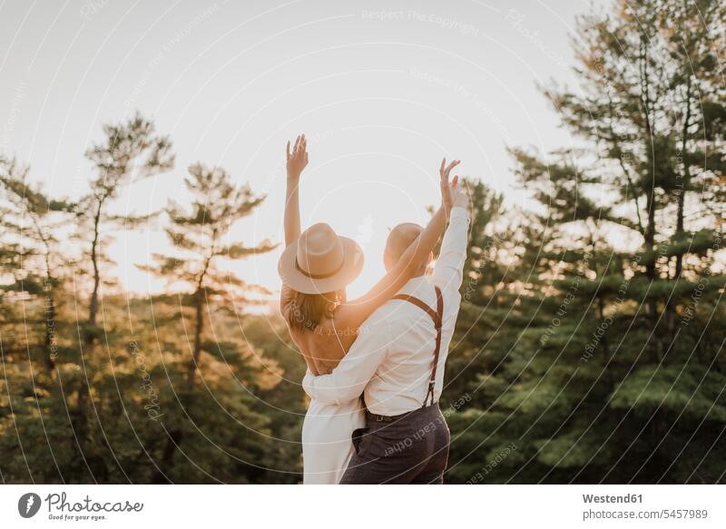 Couple with hand raised looking at sunset while standing in forest color image colour image outdoors location shots outdoor shot outdoor shots sunsets sundown