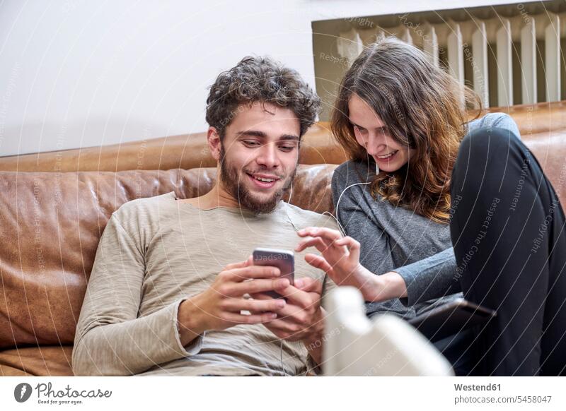 Young man and young woman sitting on couch sharing cell phone and earphones mobile phone mobiles mobile phones Cellphone cell phones share couple twosomes