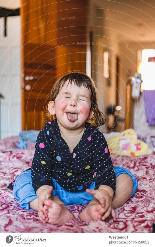 Portrait of toddler sitting barefoot on bed sticking out tongue - a ...