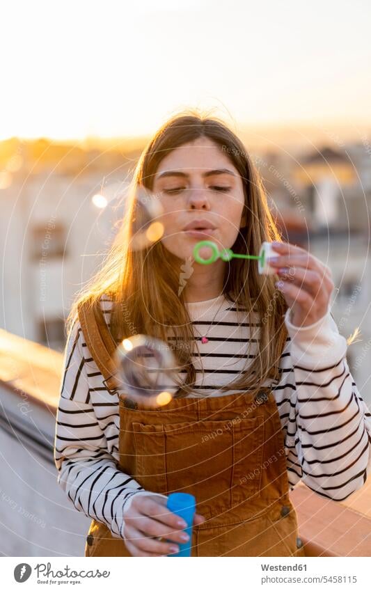 Portrait of young woman blowing soap bubbles on roof terrace in the evening females women portrait portraits deck rooftop Adults grown-ups grownups adult people