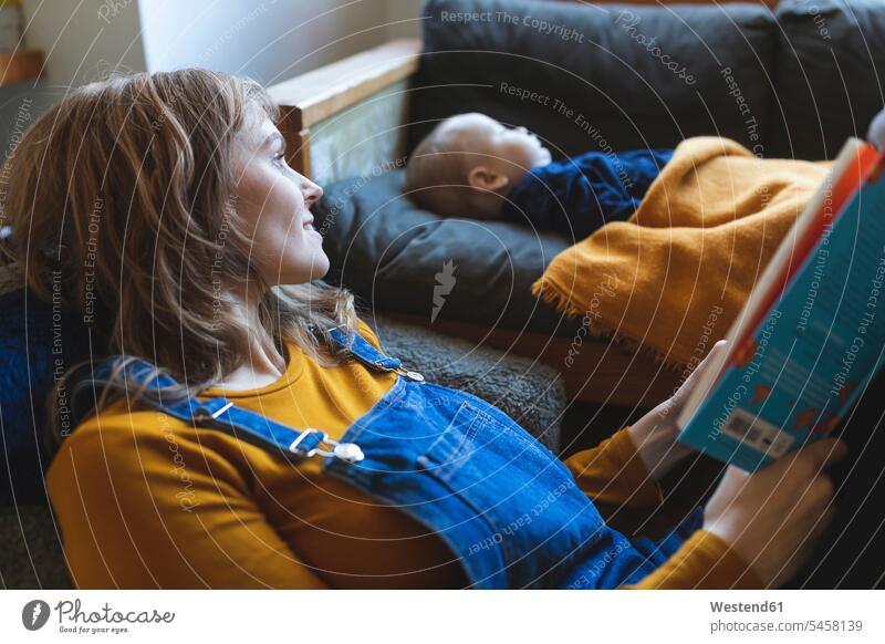 Woman reading book in living room with little son sleeping books couches settee settees sofa sofas chairs Arm Chair Arm Chairs armchairs relax relaxing smile