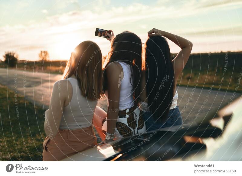 Female friends taking selfie while standing on road during sunset color image colour image outdoors location shots outdoor shot outdoor shots sunsets sundown