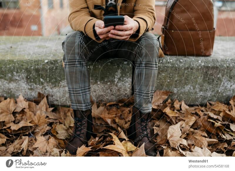 Spain, Igualada, close-up of man sitting down using cell phone surrounded by autumn leaves men males autumn leaf Seated mobile phone mobiles mobile phones