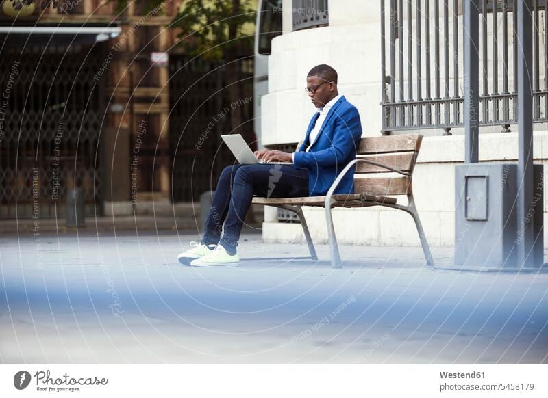 Young businessman wearing blue suit jacket sitting on bench and using laptop human human being human beings humans person persons African black black ethnicity