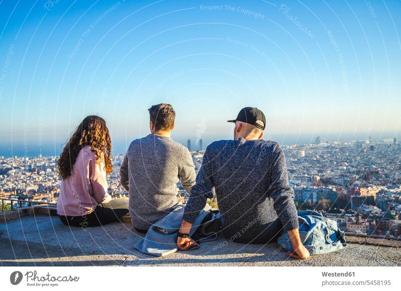Spain, Barcelona, three friends sitting on a wall overlooking the city Seated town cities towns walls mate outdoors outdoor shots location shot location shots