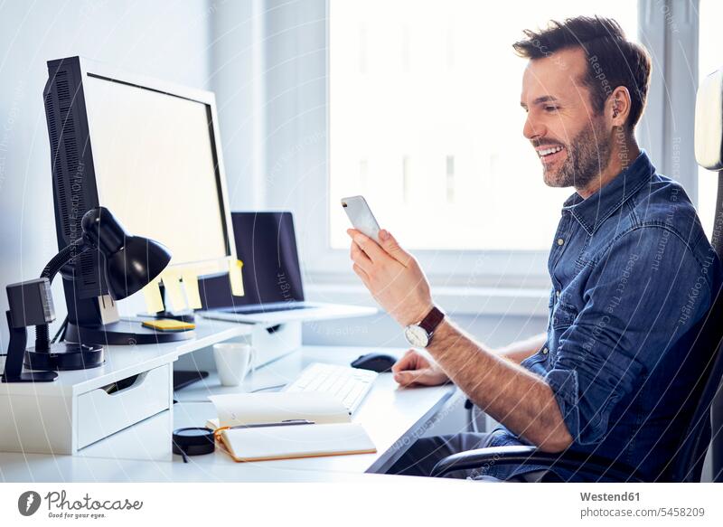 Smiling man using cell phone at desk in office desks men males offices office room office rooms mobile phone mobiles mobile phones Cellphone cell phones smiling