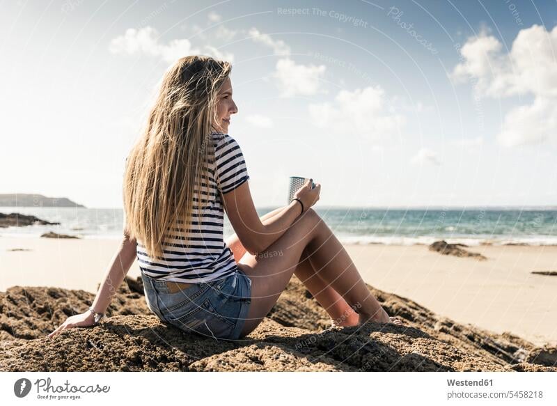 Young woman sitting in a rock at the beach, relaxing with a cup of tea drinking Seated beaches rocks Taking a Break resting break relaxation relaxed Tea Teas