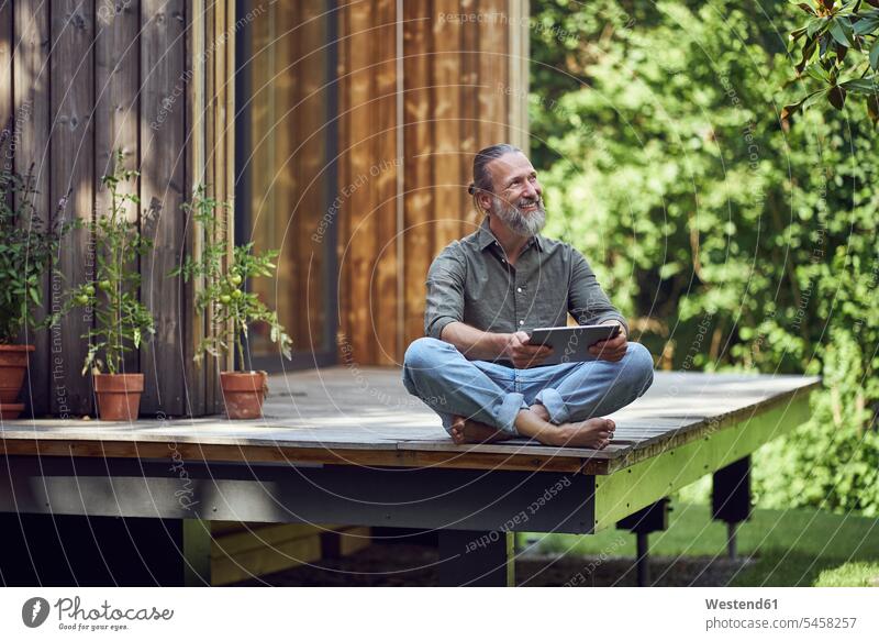 Bearded mature man holding digital tablet contemplating while sitting outside tiny house color image colour image Germany leisure activity leisure activities