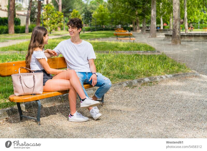 Young couple sitting on a bench and talking to each other in a park bags hand-bag hand-bags handbags benches smile Seated speak speaking summer time summertime