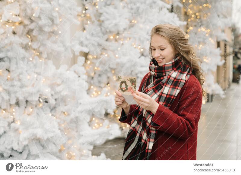 Portrait of smiling young woman standing on the street with Christmas card in front of white Christmas trees scarfs scarves smile seasons hibernal delight