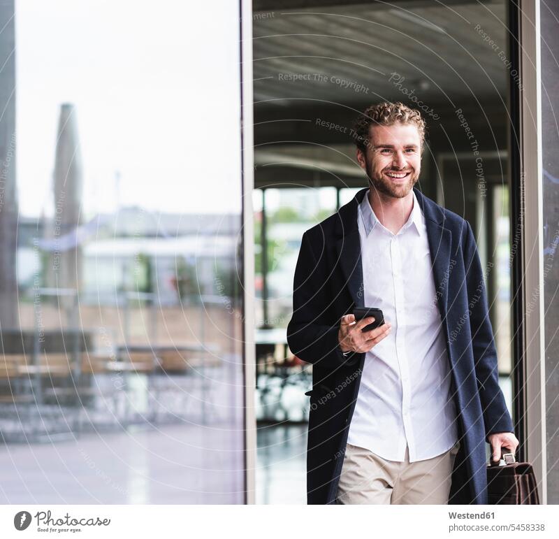 Smiling young businessman holding cell phone leaving office smiling smile Businessman Business man Businessmen Business men mobile phone mobiles mobile phones