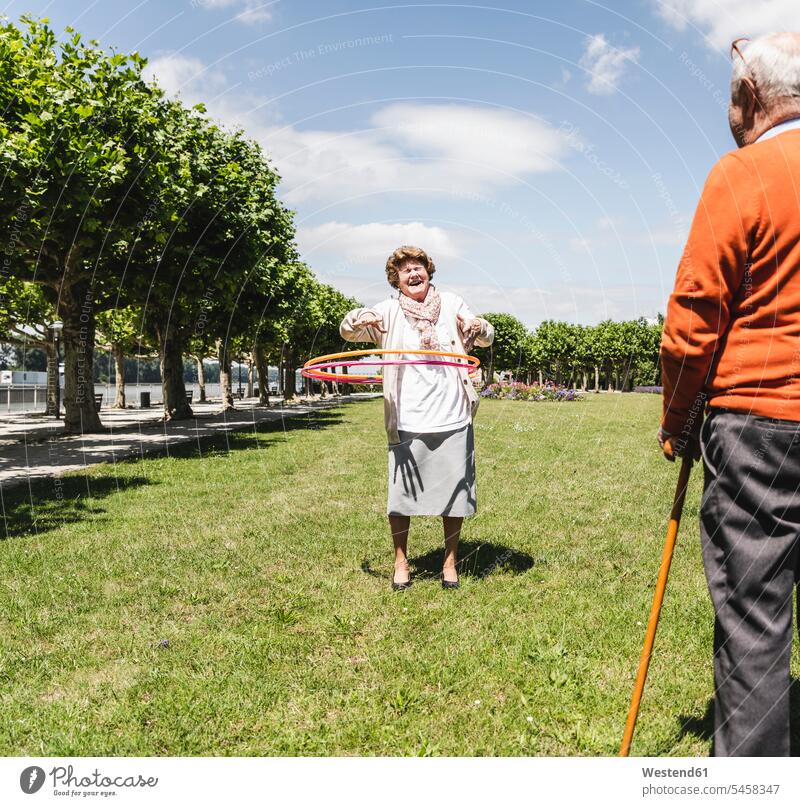 Senior watching elderly lady playing with a hoola hoop park parks summer summer time summery summertime senior couple elder couples senior couples adult couple